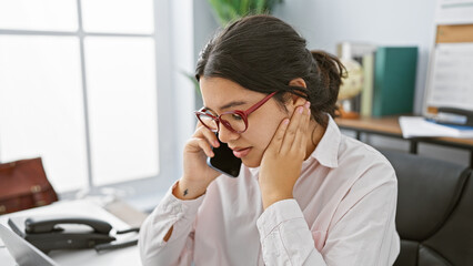 A professional young hispanic woman in glasses using a phone in a modern office setting.