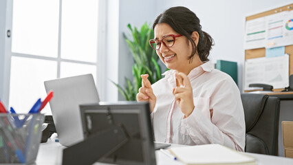 A joyful hispanic woman in an office crosses her fingers for good luck while working at a desk with a laptop and office supplies.