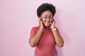 Beautiful african woman with curly hair standing over pink background covering ears with fingers...