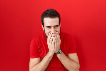 Young hispanic man wearing casual red t shirt laughing and embarrassed giggle covering mouth with...
