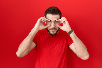 Young hispanic man wearing casual red t shirt trying to open eyes with fingers, sleepy and tired...