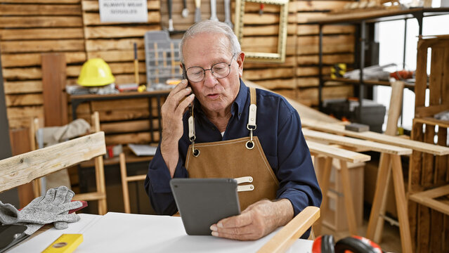 Mature carpenter having a lively chat on his smartphone, working away on his touchpad amidst the woodwork bustle of his carpentry workshop.