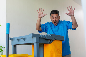 A middle-aged male janitor in a blue uniform looks surprised, raising his hands up while with his...