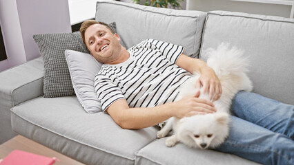 Confident young caucasian man joyfully relaxing on sofa with his happy dog, the person's cheerful...