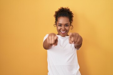 Young hispanic woman with curly hair standing over yellow background pointing to you and the camera...