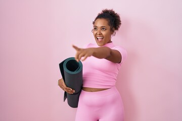 Young hispanic woman with curly hair holding yoga mat over pink background pointing with finger...
