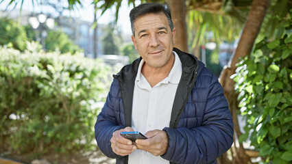 A confident mature man with a smartphone in a city park, portraying modern urban life.