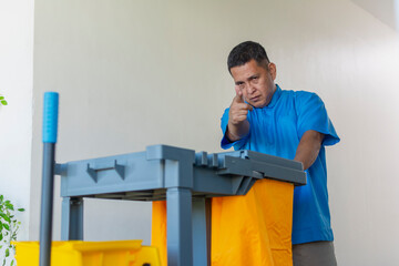 A stern looking middle-aged male janitor in a blue uniform shoots with his finger while pushing a...