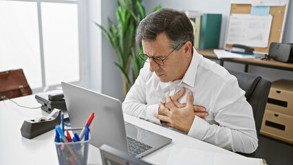 A middle-aged man in glasses experiencing discomfort at his office desk, clutching his chest in potential health distress
