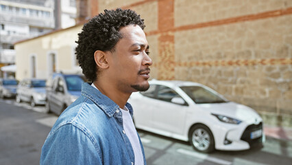 Handsome adult man with curly hair casually dressed in denim, standing on a sunny urban street.