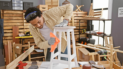 Handsome adult man with curly hair, wearing safety glasses, refinishes a stool in a carpentry...