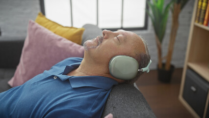 A mature man relaxes with headphones on a sofa in a cozy living room, implying serenity and leisure...