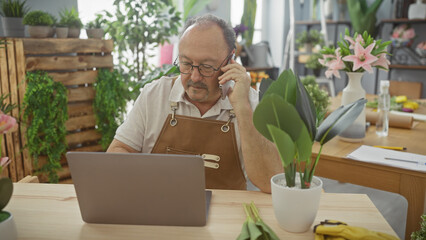 Mature man with glasses talking on phone in a flower shop surrounded by plants and using a laptop.