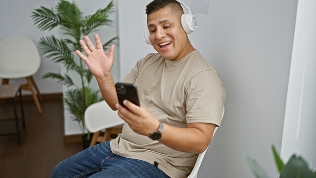 Cheerful young latin man enjoys engaging video call while sitting in a waiting room chair, embracing digital communication