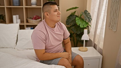 Serious young latin man sitting in cozy bed, concentrating deeply in his comfortable apartment's...