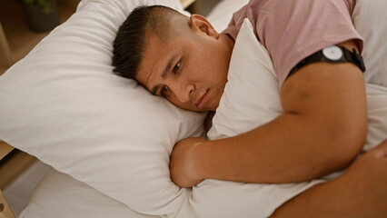 Sad young latin man hugging his pillow, laying in bed with a serious look, possibly facing morning...