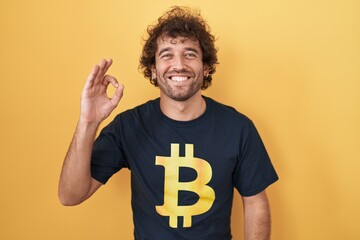 Hispanic young man wearing bitcoin t shirt smiling positive doing ok sign with hand and fingers. successful expression.