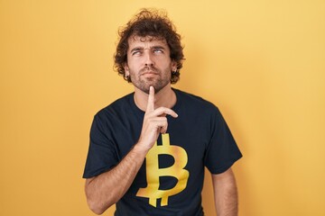 Hispanic young man wearing bitcoin t shirt thinking concentrated about doubt with finger on chin...