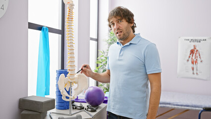 Handsome bearded man pointing at spine model in a rehabilitation clinic room with exercise...
