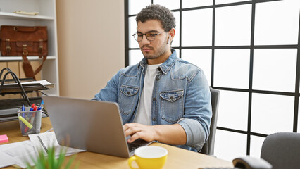 Focused man in glasses and denim typing on a laptop in a well-lit modern office room, exhibiting...