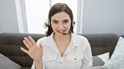 A smiling young hispanic woman wearing a headset waves in a modern living room, depicting a...