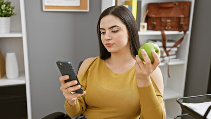 Hispanic woman multitasking with smartphone and apple in modern office setting, exuding professionalism and health-consciousness.