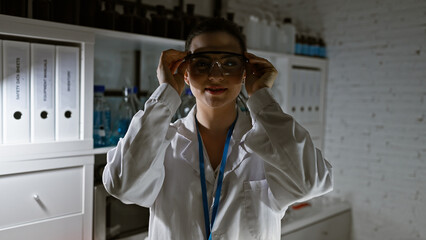 A young hispanic woman scientist in a lab wearing safety goggles and a white coat stands confidently indoors.