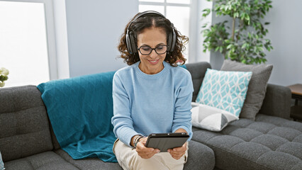 Mature hispanic woman with glasses and headphones using tablet on living room couch