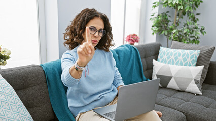 Hispanic woman with glasses gesturing indoors on a couch with a laptop, giving a stern look in a...