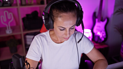 Hispanic woman with headphones in a gaming room at night, focused on a screen with neon lighting.