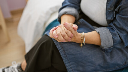 An adult woman sitting indoors, clasping hands in a thoughtful pose, suggesting reflection or contemplation.
