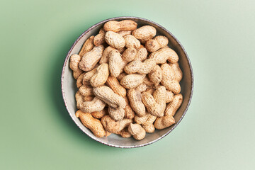 Overhead view of unshelled peanuts in a ceramic bowl on a green background, implying healthy snacking.