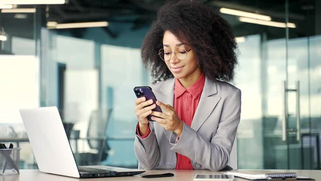 Happy young african american businesswoman in a jacket is using phone sitting at workplace in business office. Black woman reading typing a message, chatting online and looking at the camera. Portrait