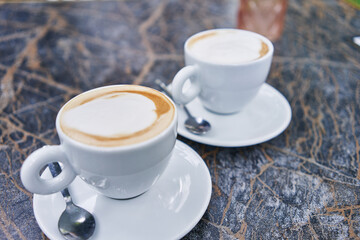 Two cups of cappuccino with frothy foam, a spoon, and saucers on a marble table.
