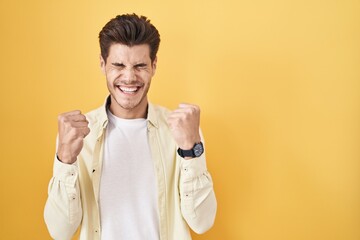 Young hispanic man standing over yellow background excited for success with arms raised and eyes closed celebrating victory smiling. winner concept.