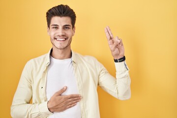 Young hispanic man standing over yellow background smiling swearing with hand on chest and fingers...