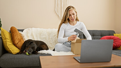 A young woman unpacking a box with her labrador dog resting on the couch in a cozy living room.