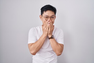 Young asian man standing over white background laughing and embarrassed giggle covering mouth with...