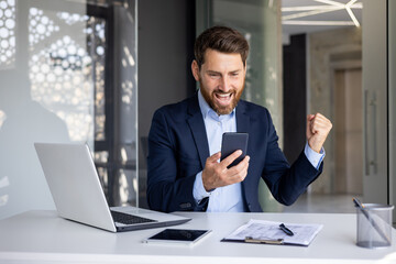 Fototapeta premium Successful and happy young businessman sitting in modern office at desk, looking at mobile phone and happy with achievement, showing victory gesture with hand