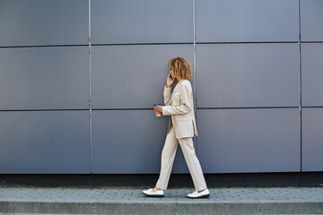 curly african american businesswoman walking with coffee and talking by phone against grey wall