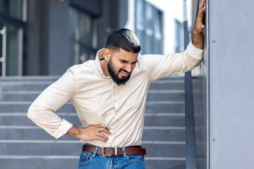 A young Muslim man in a shirt is standing outside an office building, leaning his hand on the wall and holding the side of his body, feeling severe pain in his stomach, grimacing from convulsions - obrazy, fototapety, plakaty