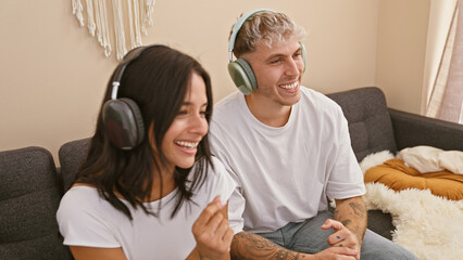 A joyful man and woman wearing headphones laugh together on a couch in a cozy living room.
