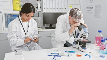 Man and woman scientists in laboratory, working with microscope and taking notes for research.