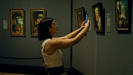 Young beautiful hispanic woman visiting art gallery taking pictures with smartphone at Art Museum...