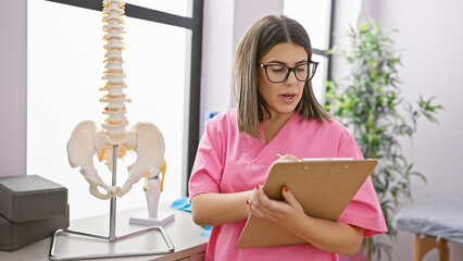 Young hispanic woman in pink scrubs reviews patient chart in clinic with spine model visible.