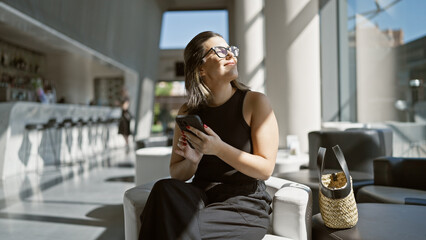 Fototapeta na wymiar Delightful hispanic woman sitting in a modern hotel hall, wearing glasses, using her smartphone, smiling while looking off to the side