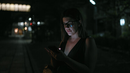 At night, beautiful hispanic woman wearing glasses holds a deep conversation over text, engrossed...