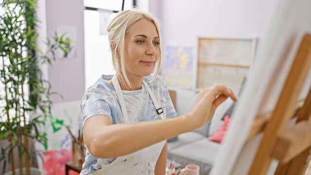 Happy young blonde woman artist, confident and smiling, enjoying her art lesson indoors at university art studio, while drawing beautiful canvas with paintbrushes