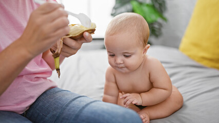 Loving mother sitting on bed, sharing a healthy banana snack with her daughter, binding their...