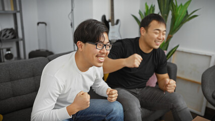 Two asian men excitedly celebrating while seated on a couch in a cozy living room environment.